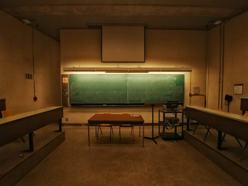 Empty teaching room with a green chalkboard, an unused lectern and rows of desks, photographed in low light